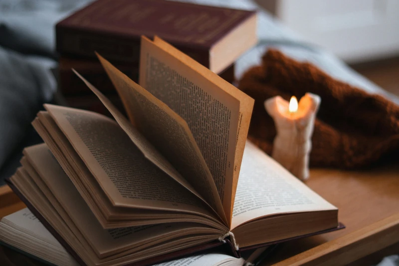 Aufgeschlagenes Buch mit sich umblätternden Seiten neben einer brennenden Kerze und einer kuscheligen Decke auf einem Holztisch, gemütliche Abendstimmung zum Lesen vor dem Schlafengehen