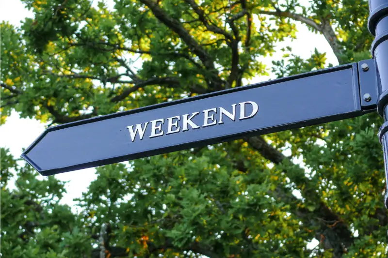 Schwarzes Straßenschild in Pfeilform mit der Aufschrift Weekend grüner Baum im Hintergrund