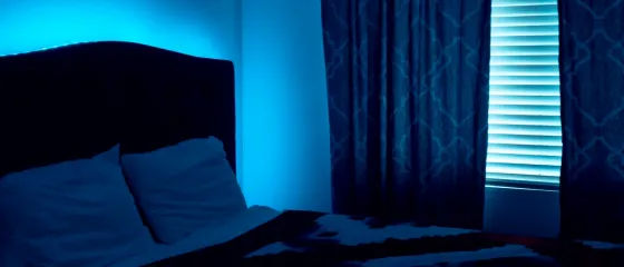 Schlafzimmer mit Doppelbett, im Hintergrund blaues LED-Ambientelicht hinter dem Bettkopfteil und an einem beleuchteten Fensterrollo