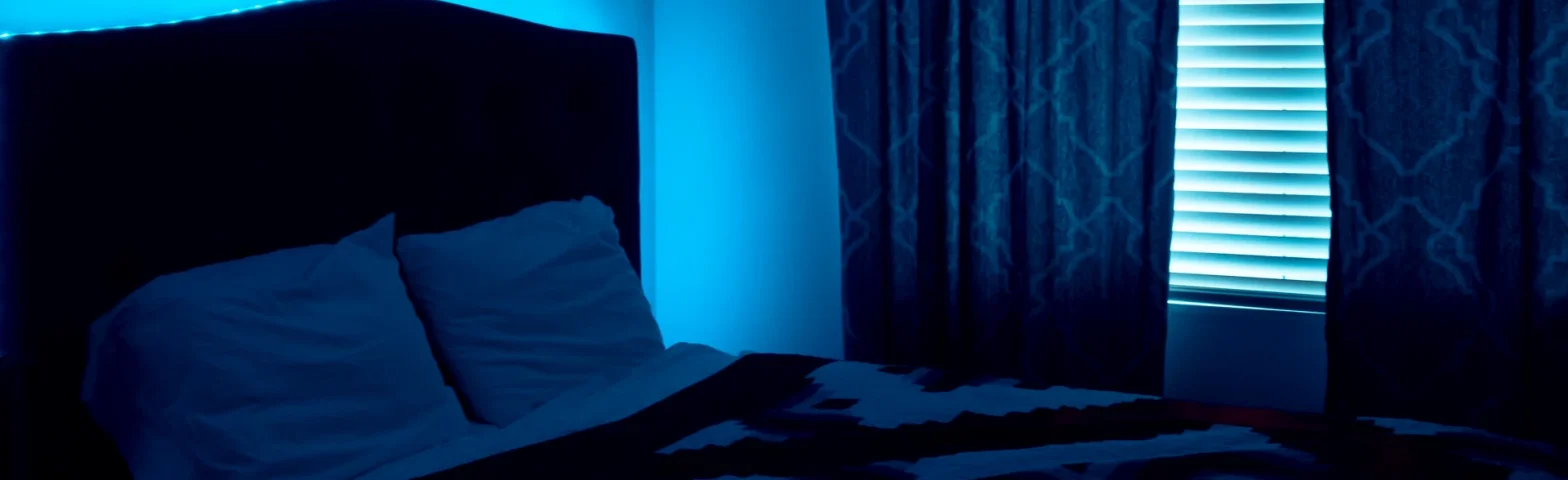 Schlafzimmer mit Doppelbett, im Hintergrund blaues LED-Ambientelicht hinter dem Bettkopfteil und an einem beleuchteten Fensterrollo