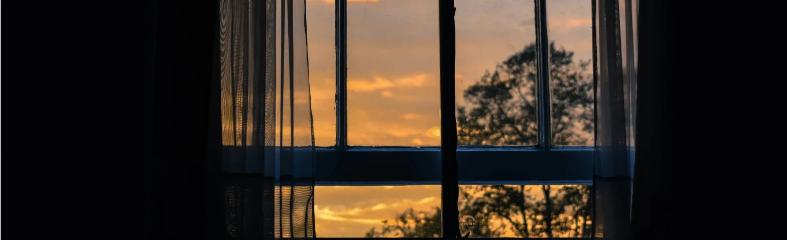 Blick durch ein Fenster mit Vorhang auf Abendhimmel und Bäume in der Dämmerung