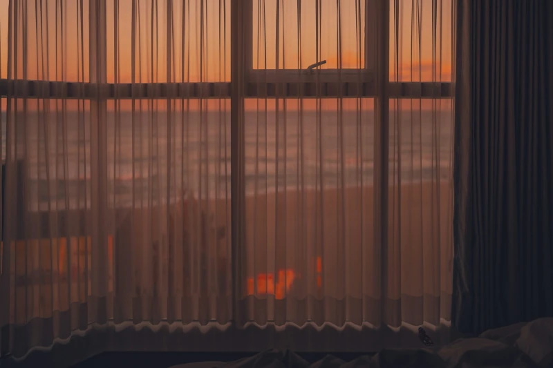 Durchscheinender Vorhang vor einem Fenster mit Blick auf Meer und Strand bei Sonnenuntergang, warmes orangefarbenes Licht
