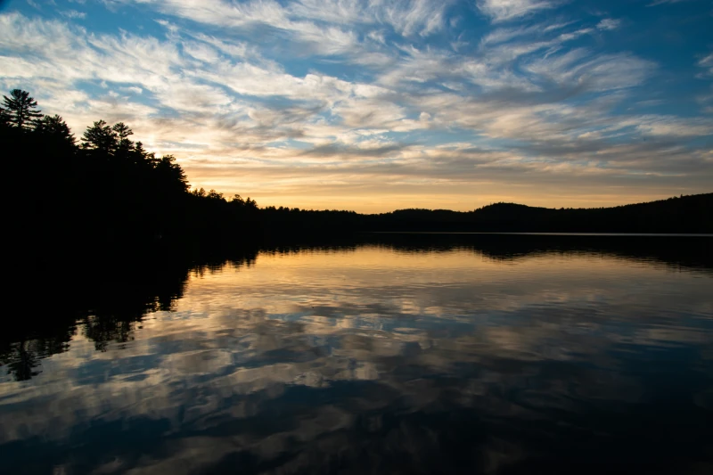 Ruhiger See bei Sonnenuntergang mit Wolkenspiegelung im Wasser und Baumsilhouetten am Ufer