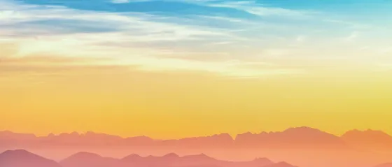 Bergpanorama in Pastelltönen bei Sonnenuntergang, Silhouetten mehrerer Bergketten vor gelb-orange-blauem Himmel