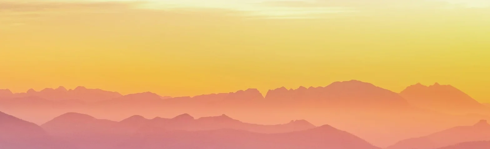 Bergpanorama in Pastelltönen bei Sonnenuntergang, Silhouetten mehrerer Bergketten vor gelb-orange-blauem Himmel